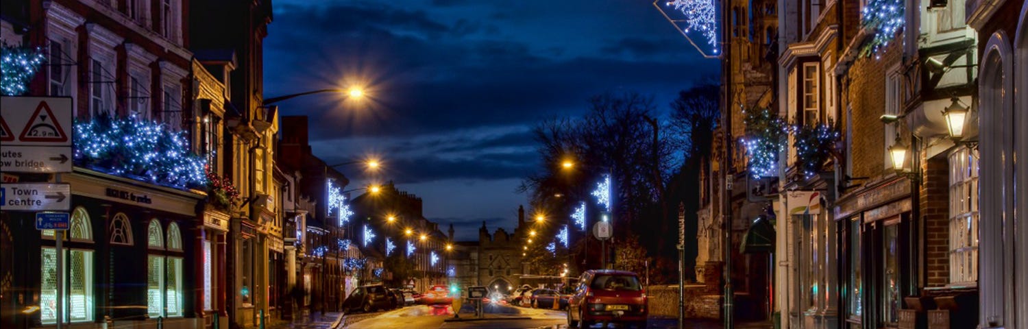 Image of a night time street scene in an English town where the road and paths are damp from evening rain and capture the reflections of street lights