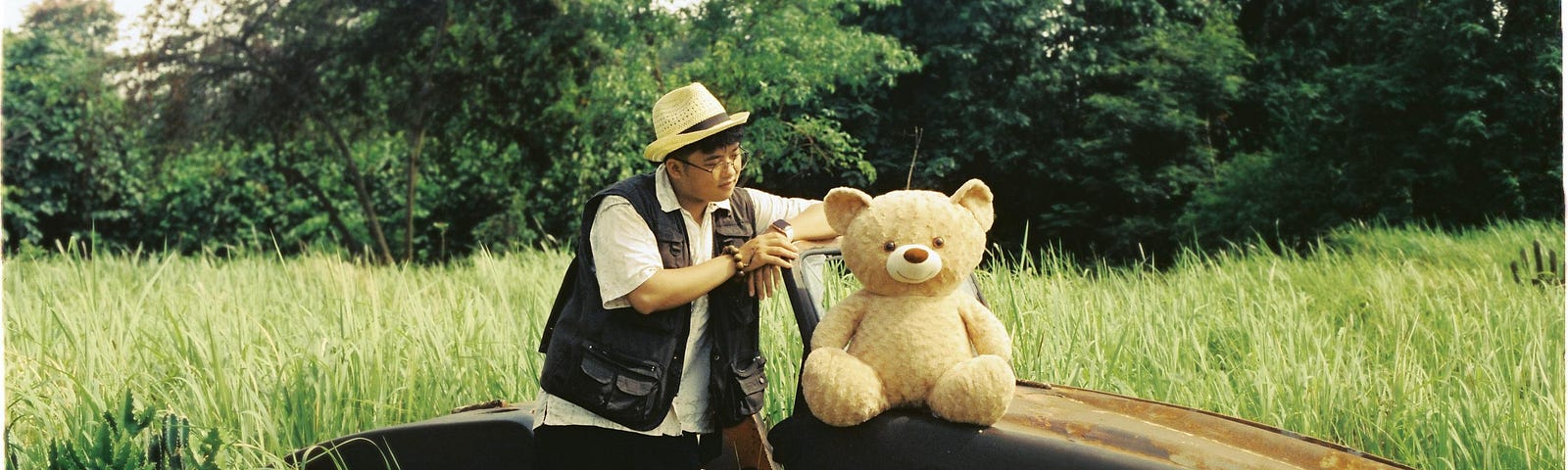 A man in a hat stands next to a teddy bear by a car wreckage on the grassland