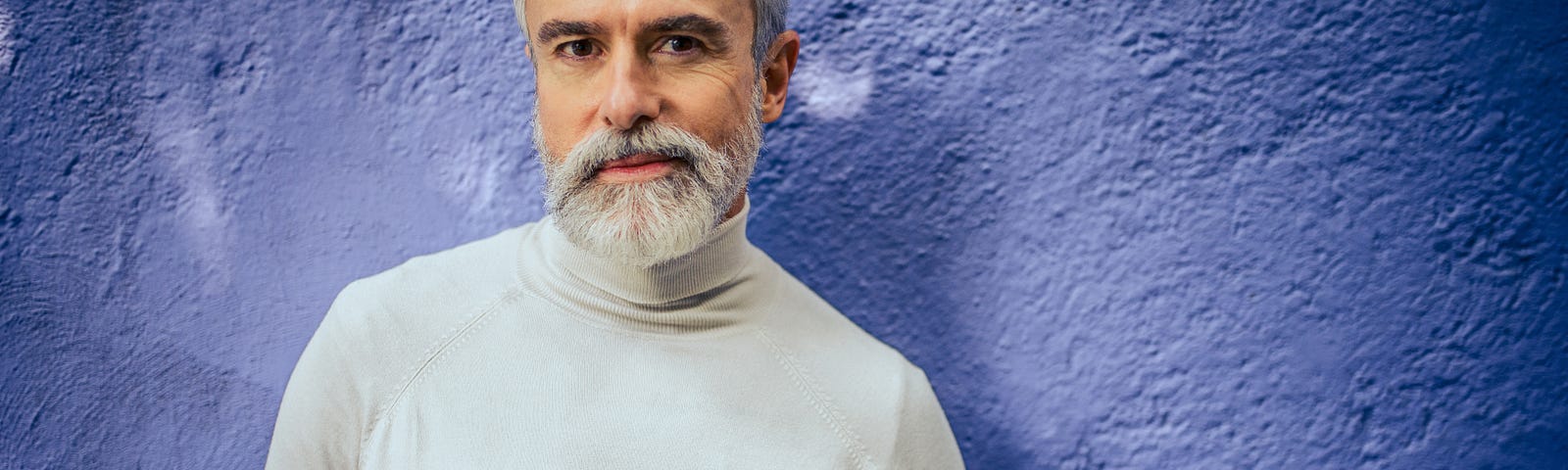 Portrait of a smiling, bearded, mature man standing in front of a blue wall.