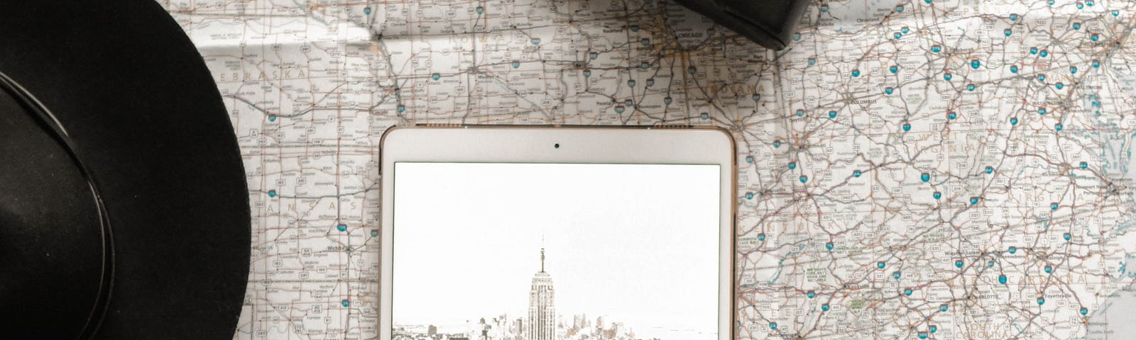 A tablet showing an image of New York, sat on a map. A hat and camera lay on the table too.