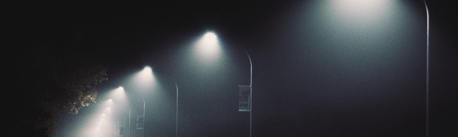 Several streetlights shine on a dark road, with one figure walking at the end.