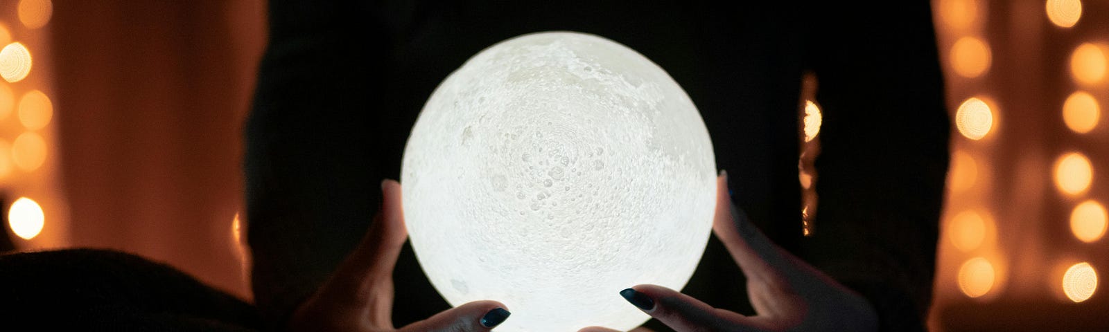 A woman’s dark, shadowy figure sits, holding a white crystal ball that looks like a little moon in her hands. It’s a body shot; you can’t see her head. Orange, brownish lights twinkle in the background.