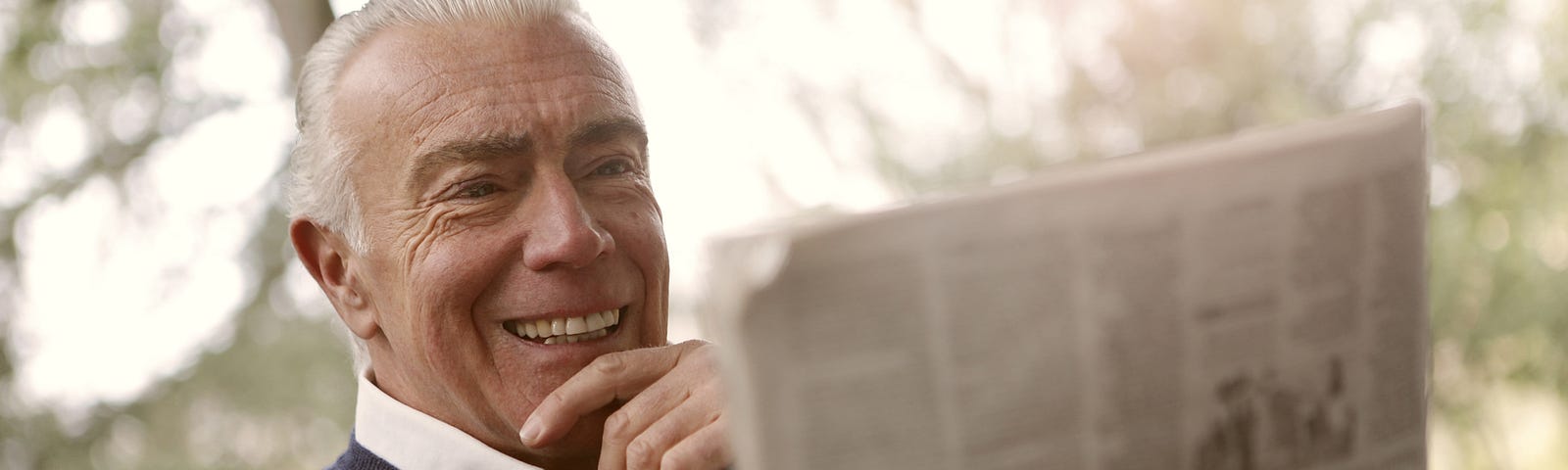 Man with white hair smiling while reading the newspaper.