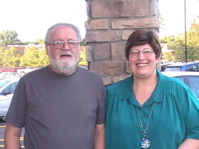 Bob, a white haired man in his 70s with glasses and a beard is on the left, and Charmaine, a woman in her 70s with short dark hair parted on the left, is on the right. She is wearing a large button down teal shirt and a long hanging necklace..