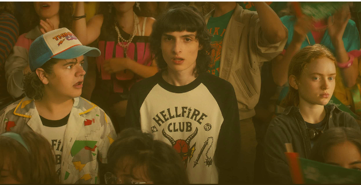 3 characters from the series, Dustin, Mike, and Max, in a school prep rally. Dustin and Mike wear Hellfire Club t-shirts.