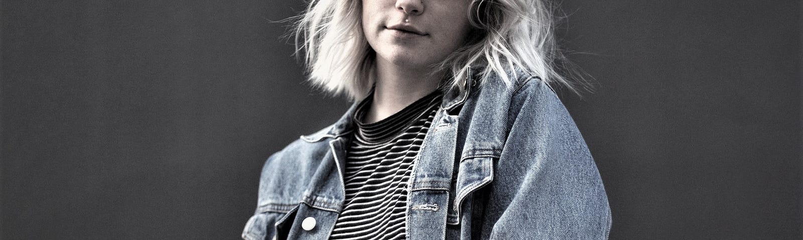 pensive girl with blonde hair wearing denim jacket and striped shirt against dark background
