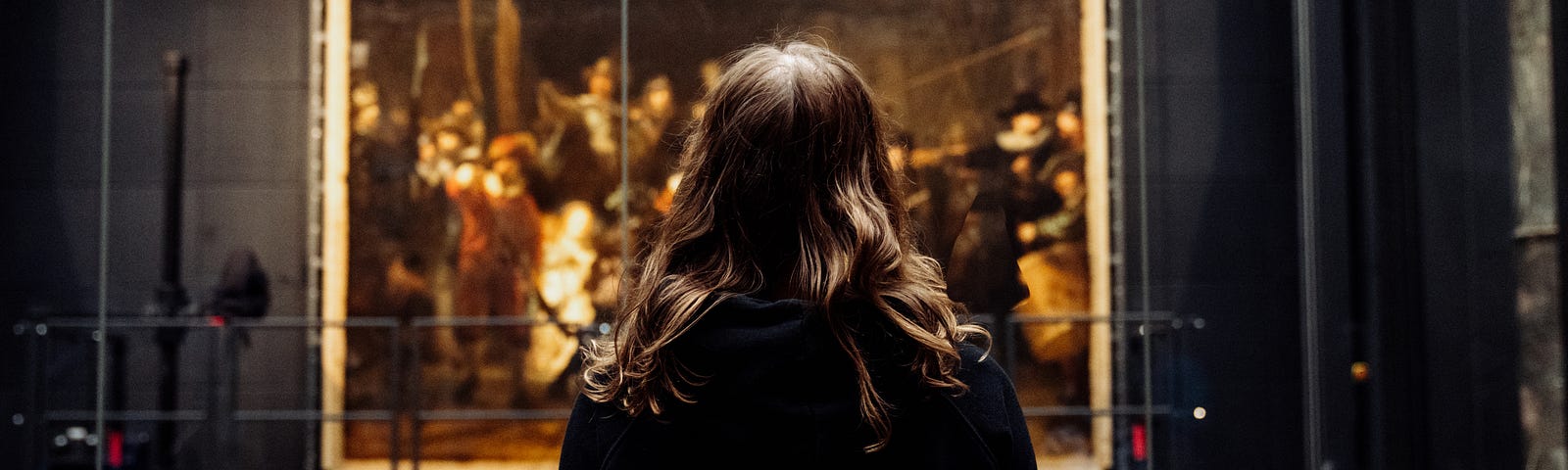 The picture features a woman’s back. She wears a black hoodie that features the letters DTNS on the back in white. She is looking at a Rembrandt painting.