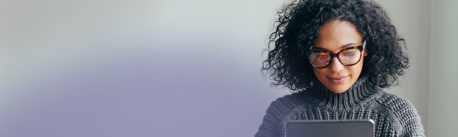 A woman with curly hair, wearing glasses and a cozy sweater, sits in a quiet workspace looking at an iPad.