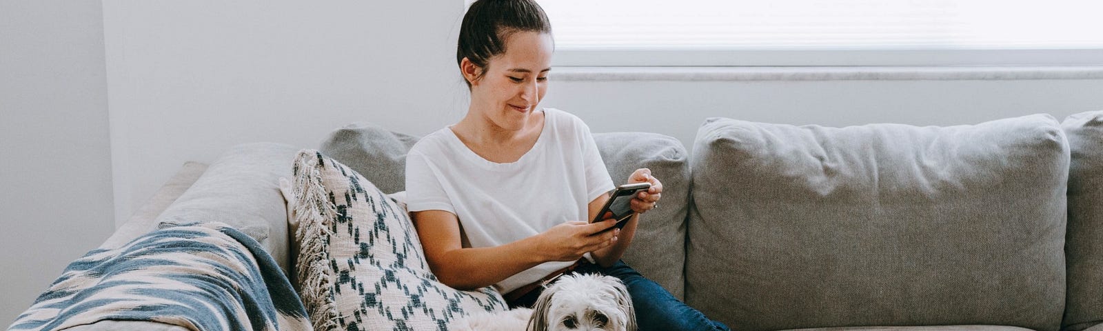 A woman smiles while looking at her phone and sitting next to her dog on the couch.