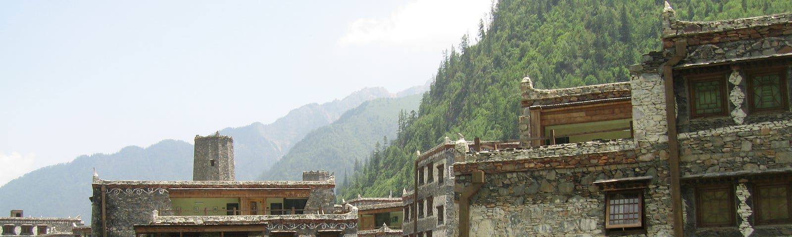 Buildings made of rocks with brown wooden trims on a green hill with mountain ranges in the background.