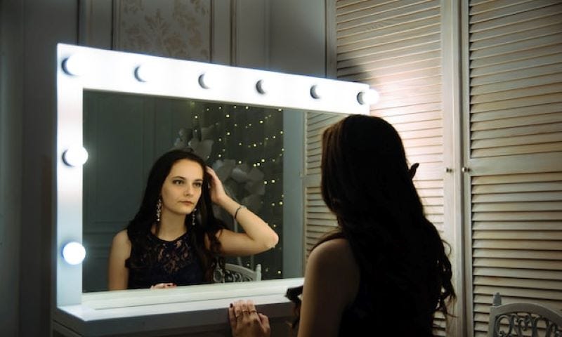 A white woman with dark hair and wearing a dark dress looking into a vanity mirror