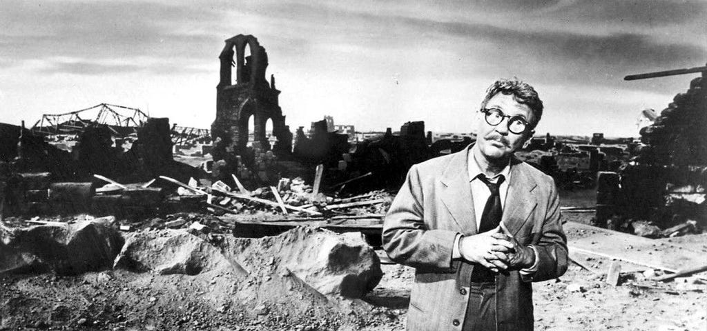 black & white image of a man wearing a suit and tie with glasses standing in a wasteland amongst rubble after a bomb went off