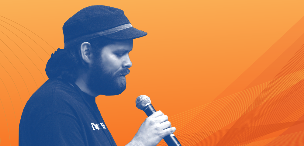 Grey-tone photo of Kin Lane wearing a hat and talking into a mic against an orange background.