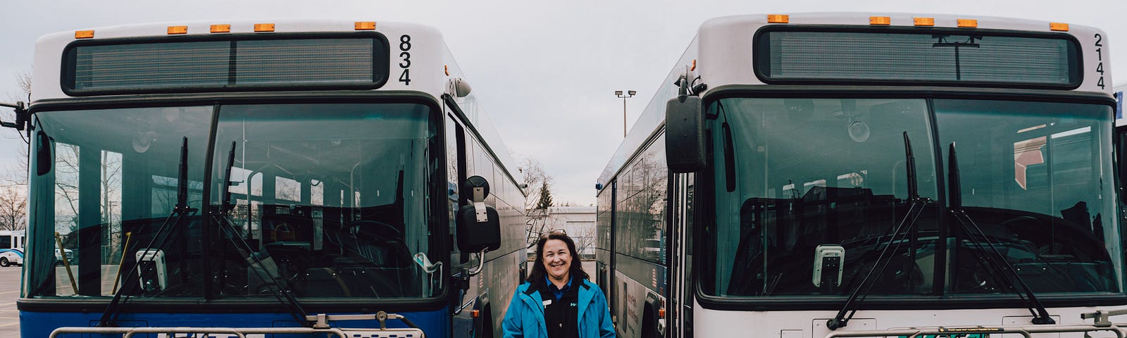 Shannon Carlton, wearing a blue rain jacket stands just off a curb in between two parked busses.