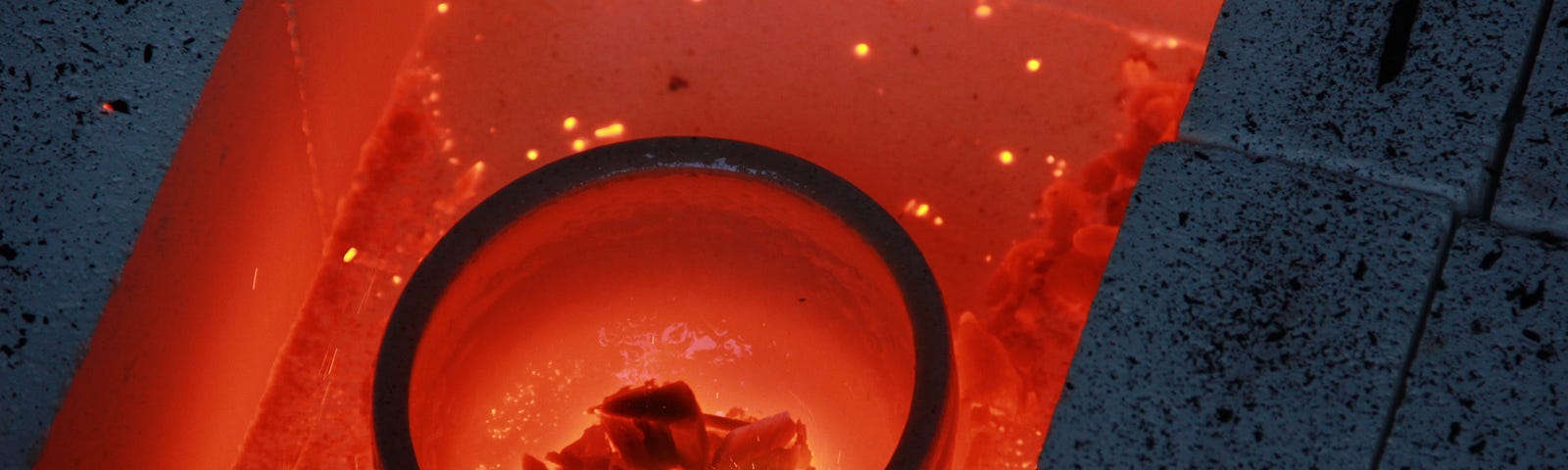 Picture of a small bowl sitting in the red hot fire in a hole surrounded by grey bricks . There are red embers in the bowl.