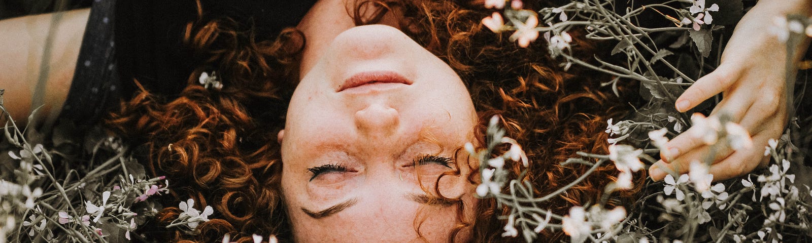 Red-haired White woman’s gently smiling face turned up-side-down on a bed of small white flowers.