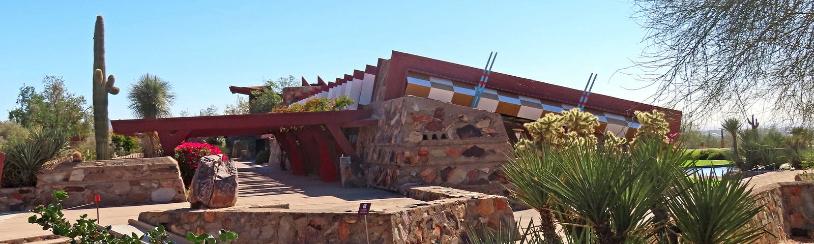 Desert brush in the foreground with a low rock wall between the garden and a large beige concrete pad. There is a low rocks ledge in a square shape with a large rock in the corner, a saguaro cactus is in the background beside a stone faced one-level sloped-roof building with a red metal portico