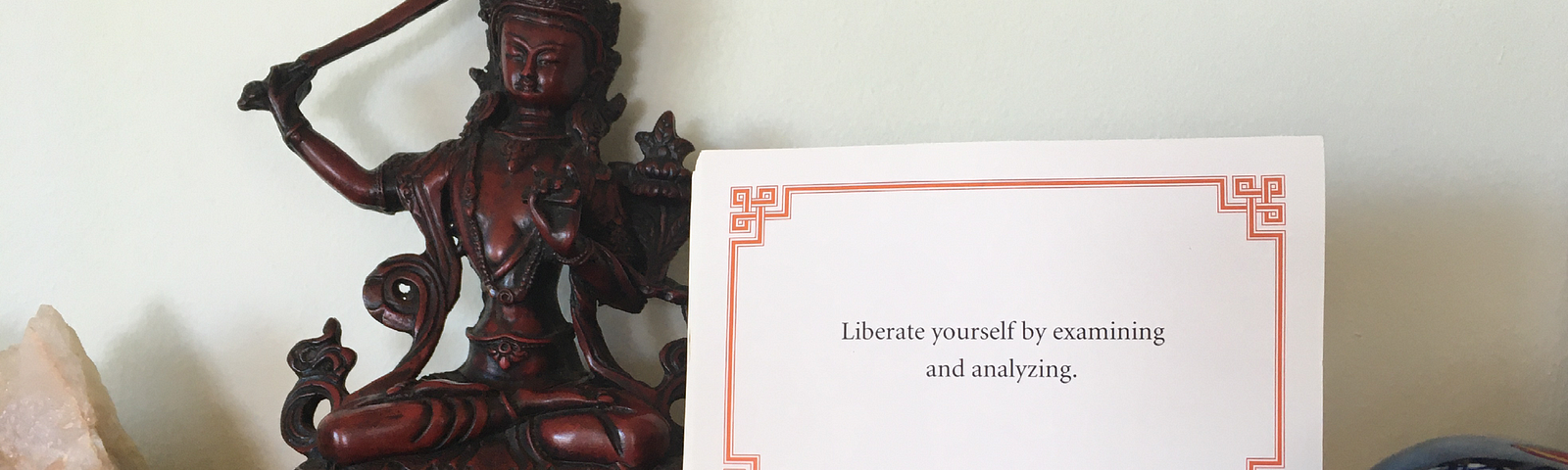 A Lojong card that reads “Liberate yourself by examining and analyzing” sits on a wooden shrine, to the right of a statue of the Bodhisattva Manjushri.