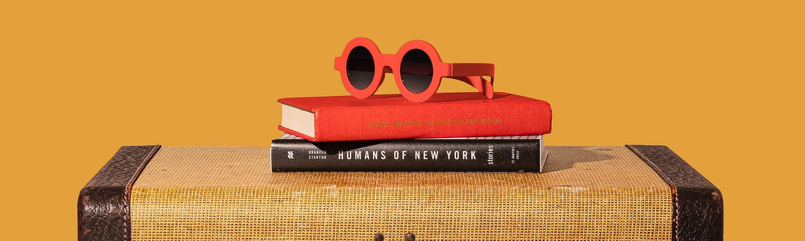 Vintage style rectangular brown suitcase with two books and round red framed sunglasses sitting on top, with orange background