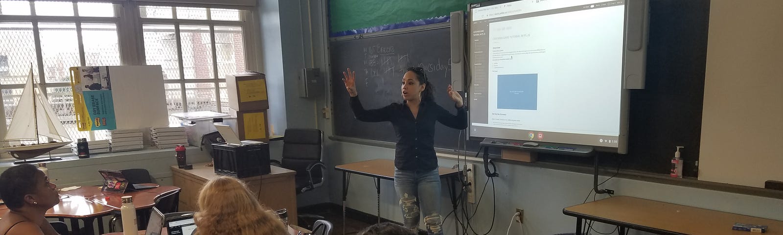 Four people in a classroom. The teacher, a woman in with dark hair, has both her arms raised and is speaking at the front.