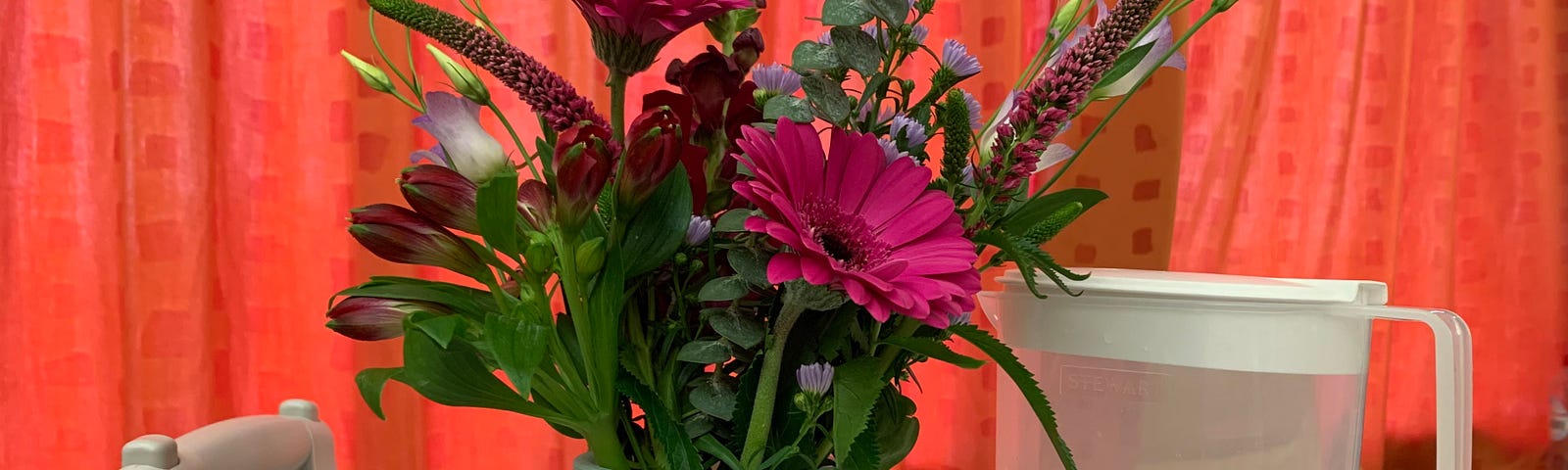 This is a photo of pink flowers in a blue vase. Thee is a plastic pitcher of water nearby with a glass next to it. The vase and water are sitting on a white table that is at the foot of a hospital bed. There are orange curtains behind the table.