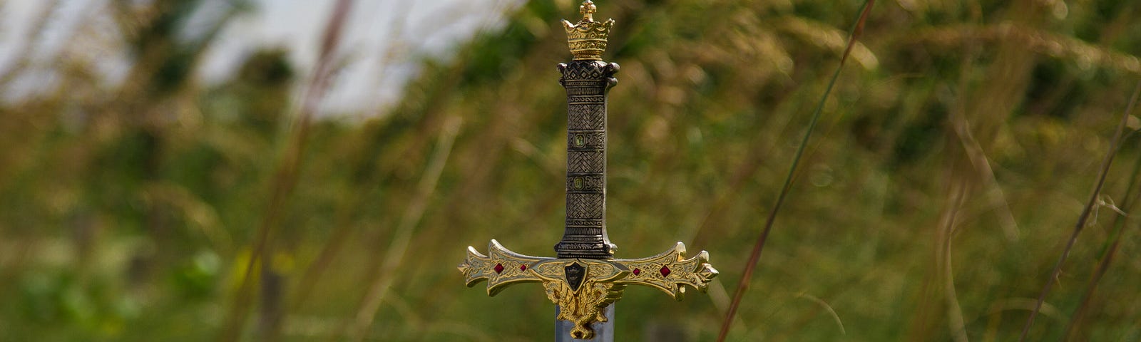 An ornate sword is stuck into the ground with a grassy field in the background.