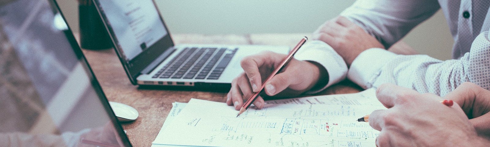 Two people writing on a piece of paper and two laptops on the table.