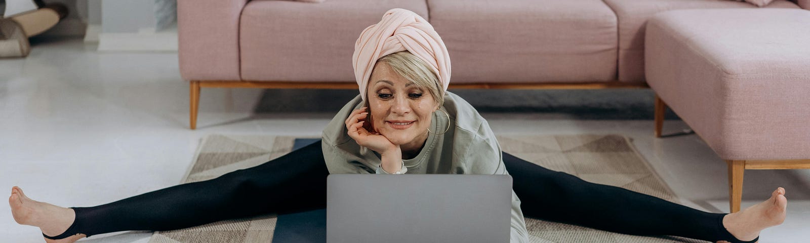 A senior woman since on the carpet in front of a pink lounge she is doing the splits while looking at her computer. She has a pink scarf on her head.