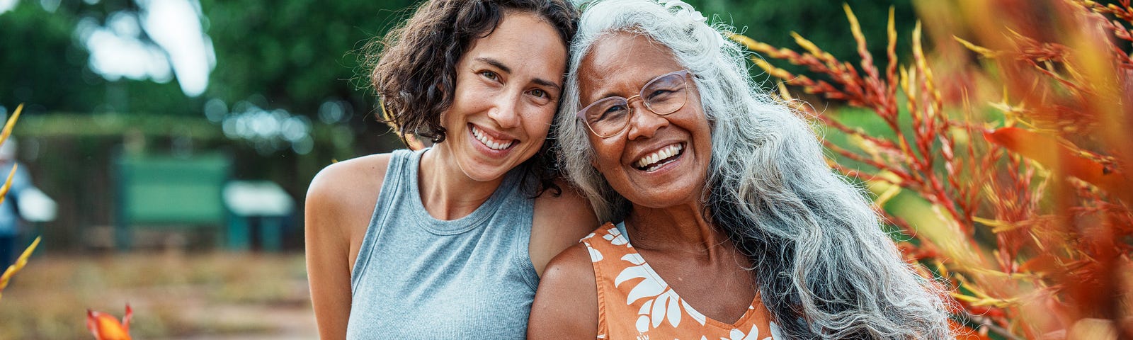 Portrait of a beautiful and healthy senior woman of Hawaiian and Chinese descent and her Eurasian adult daughter smiling directly at the camera while sitting together outside, surrounded by orange and yellow tropical foliage in Hawaii.