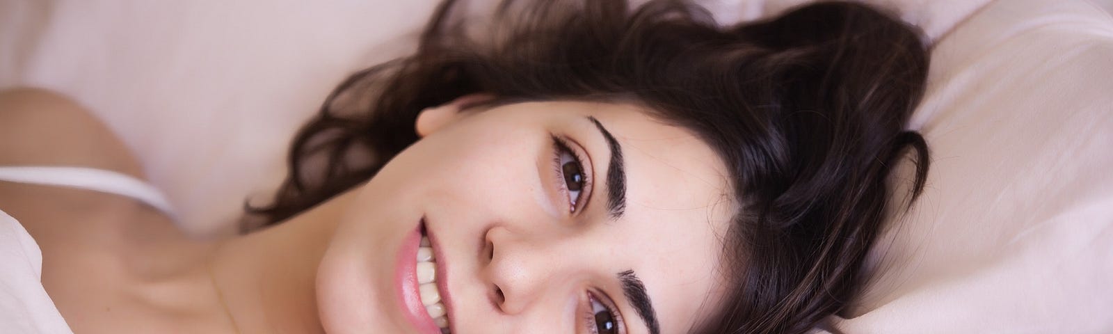 Woman with her head on a white pillow, smiling and looking alert. She has brown hair and eyes and light skin