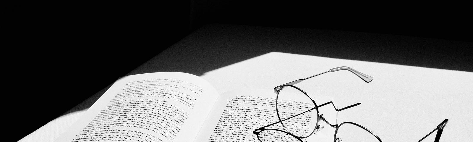 A black and white photo shows an open book lying flat on a surface bathed in sunlight. A pair of eyeglasses rests at an angle on part of the open book.