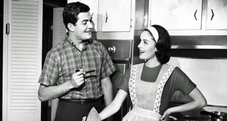 A black and white photo of a young married couple in the kitchen, with the woman wearing an apron and holding an iron.