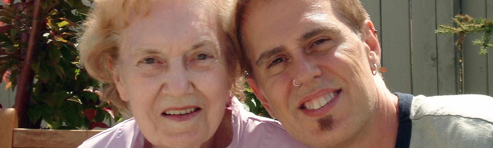 Elderly woman looks into the camera — young man has his arm around her smiling.