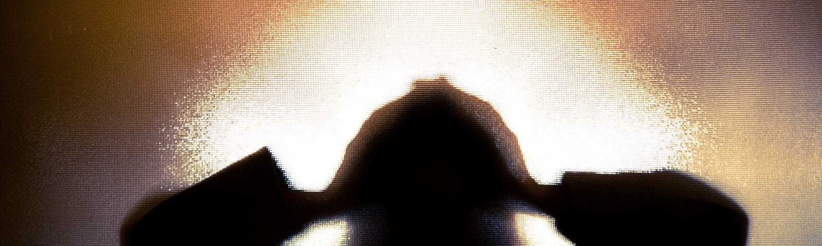 Colour backlit image of the silhouette of a woman with her hands on her head in a gesture of despair. The silhouette is distorted, and the arms elongated, giving an alien-like quality. The image is sinister and foreboding, with an element of horror. It is as if the ‘woman’ is trying to escape from behind the glass. Horizontal image with copy space.