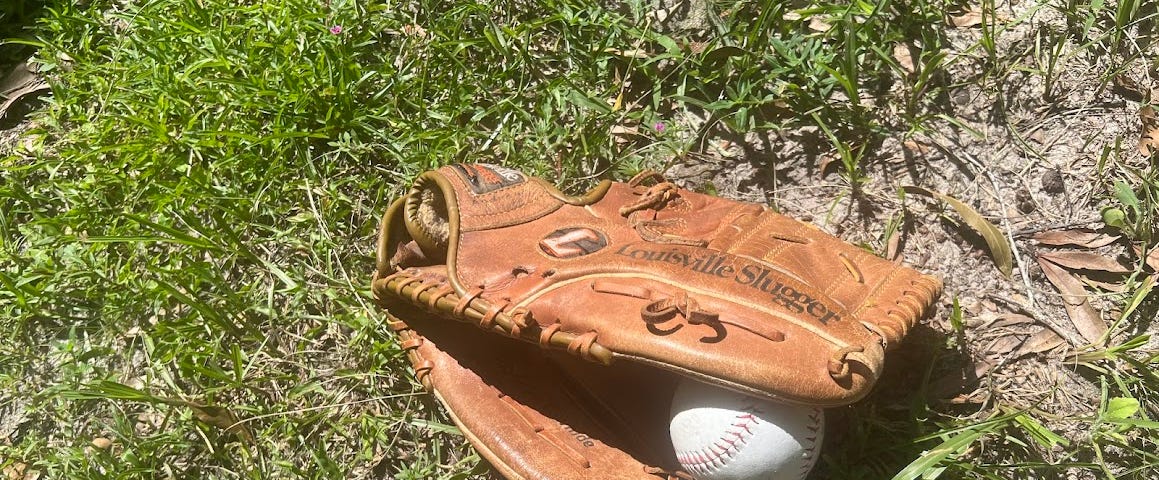 Photo of an old Louisville Slugger baseball glove lying on the ground which is a mixture of dirt and grass. The glove has a white baseball in it that is halfway visible.