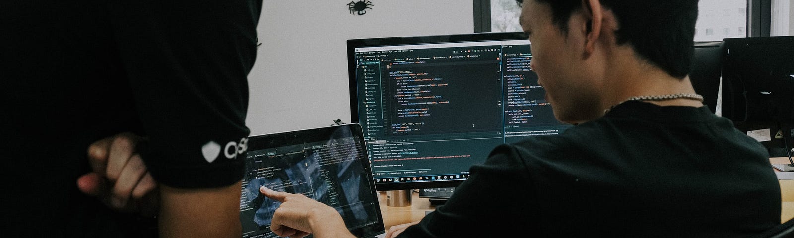 A man is pointing to a laptop screen, while another person is looking at it over their shoulder. The screen has code written on it.