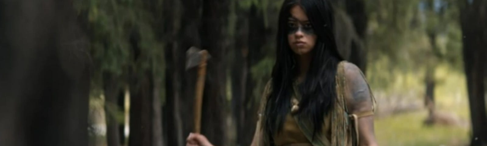 A young indigenous woman with long black hair and wearing buckskin stands in a forest in daylight. She has black face paint across her eyes and holds a tomahawk in her right hand.