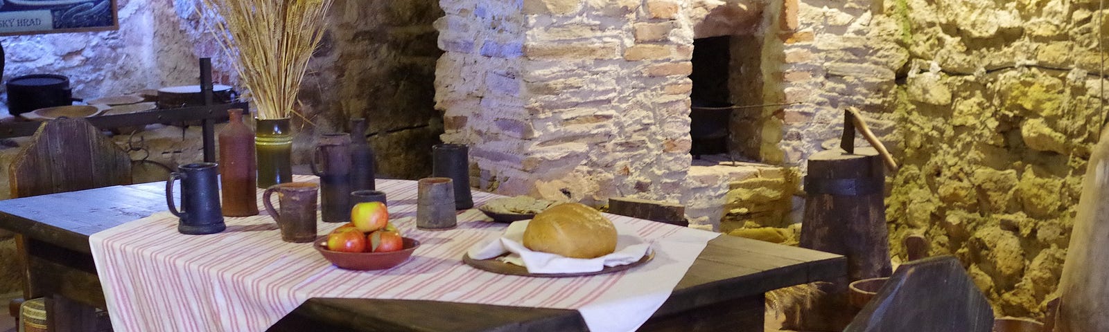 Simple wood table with a white table clothe set diagonally, a round loaf of bread, pottery cups, and a bowl of apples. The table sits in front of a corner fireplace.