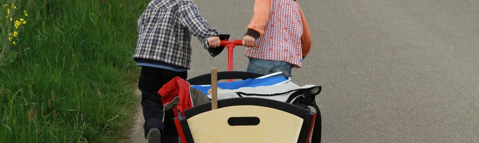 Two young children pulling a wagon filled with something flat. One of the children has a brown hard hat that covers the ears.