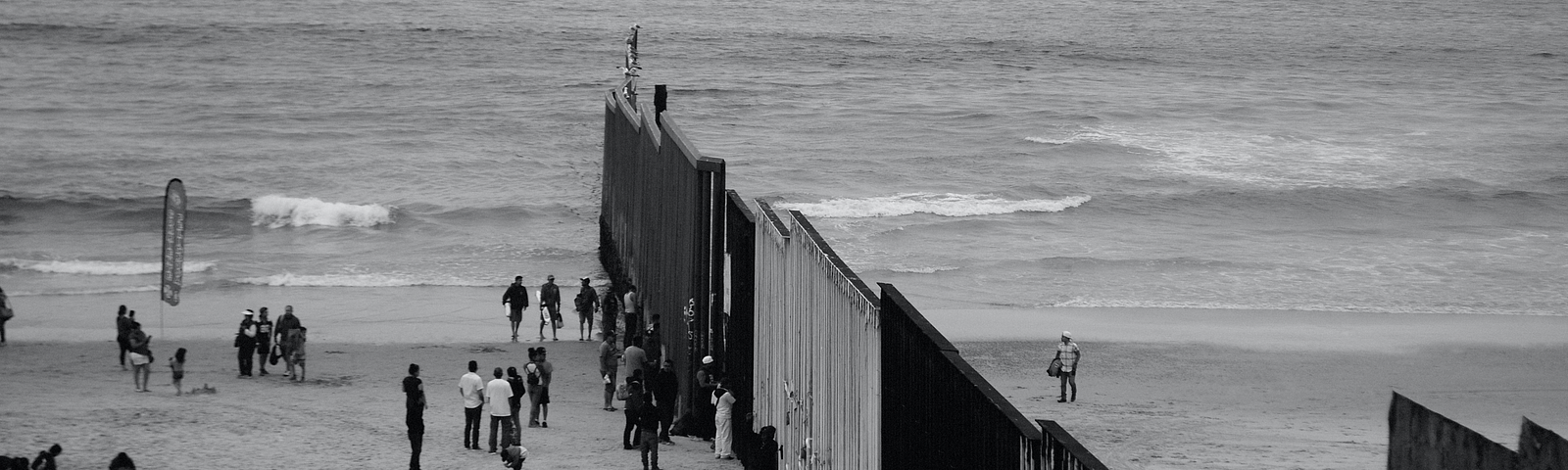 Black-and-white photograph of people on a beach, divided by a tall fence.