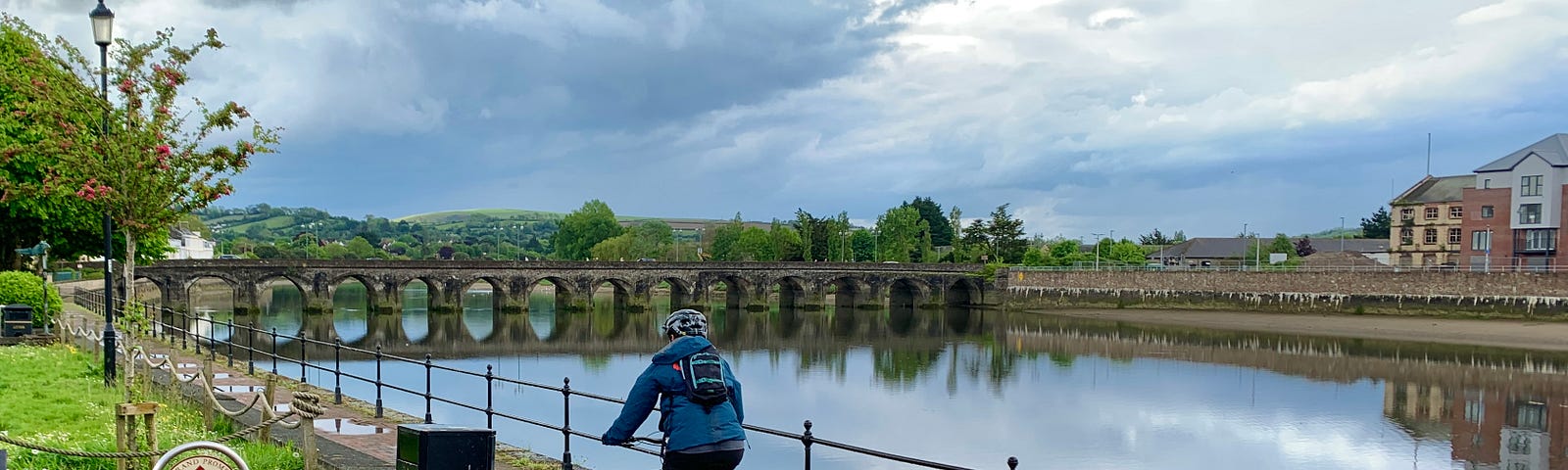 A person riding a bike along the side of a river with a bridge in the background.