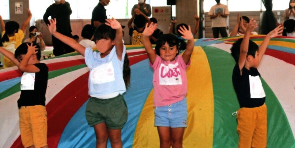 Five and six year-old children with their hands raised high, standing on a rainbow-striped circle cloth that is ballooning up behind them