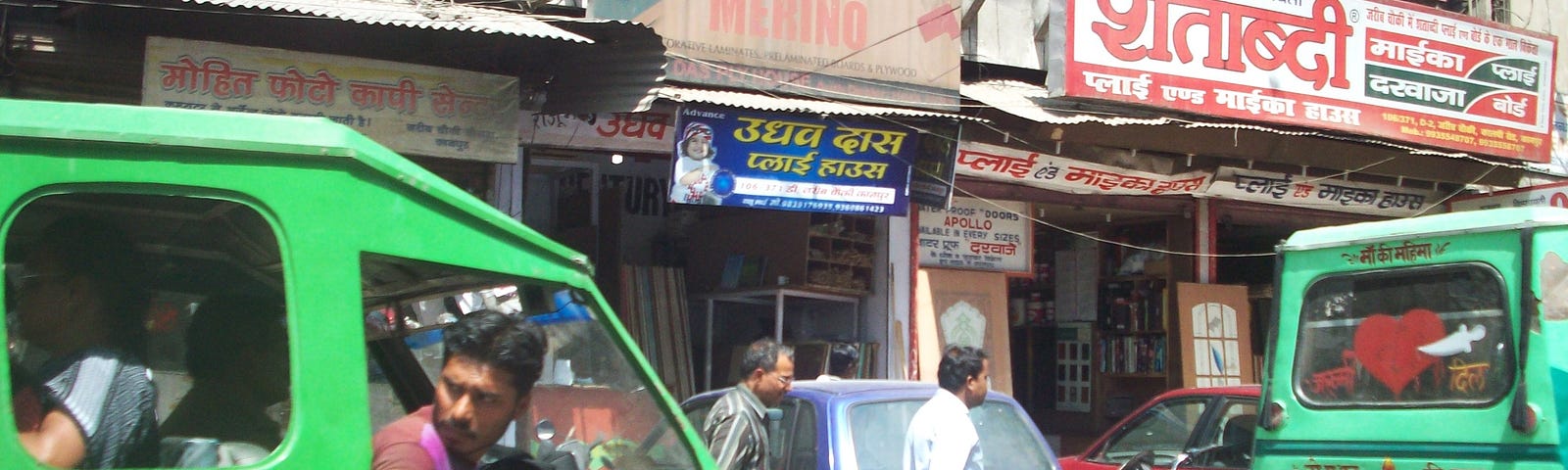 A street scene in Uttar Pradesh, India. A green van is stuck in traffic, and the driver is looking behind. Two men are passing by.