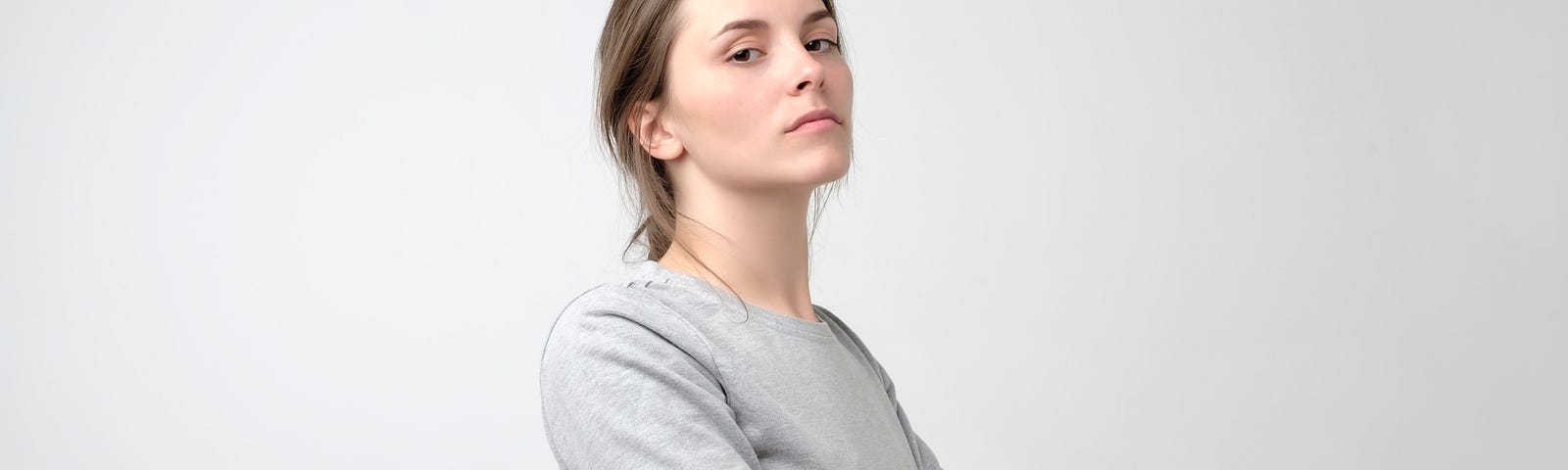 Young woman standing looking confident or arrogant in front of gray background, crossing her arms across her chest.