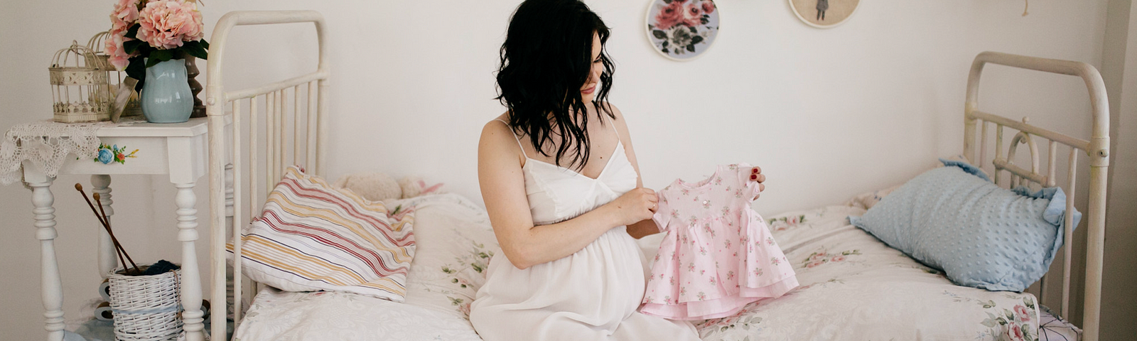 Pregnant woman in a white sun dress sitting on a child’s bed folding a tiny pink dress.