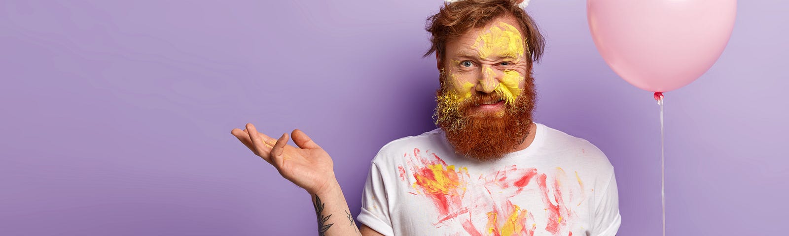 A man with red hair, a beard and tattoos on his arms, wears a unicorn hat. He has red and yellow paint covering his face and shirt. He holds his right hand up and has a somewhat puzzled expression on his face, all while holding a pink balloon in his left hand. The background behind him is purple.