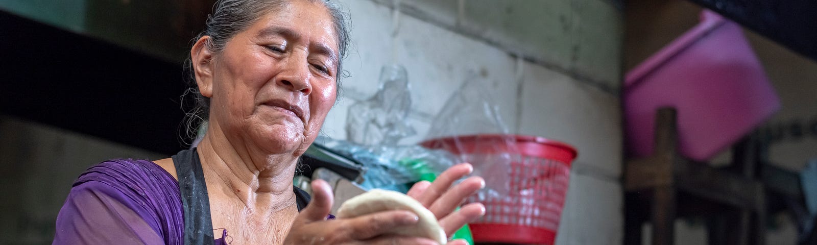 Woman in an apron shaping dough in her hands in a small store with concrete walls.