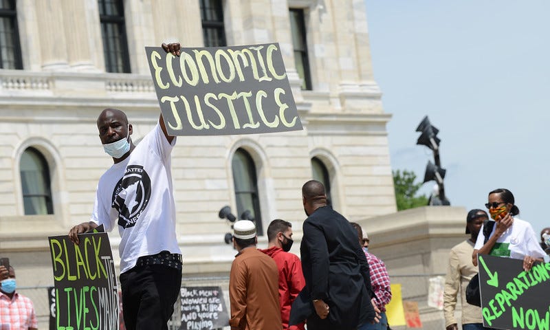 A group of Black Americans demonstrate outside a government building. They carry signs that read, “Economic Justice,” “Black Lives Matter,” and “Reparations Now.”