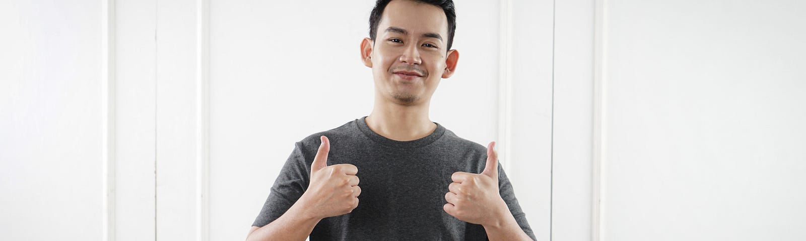 A young man in a black t shirt with his thumbs up on both hands, smiling against a backdrop of white doors