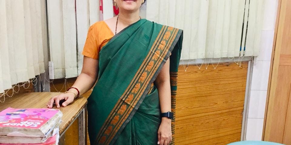 A woman in a sari, a traditional garment of India, stands in a dental clinic. A dentist’s chair is visible to her left.